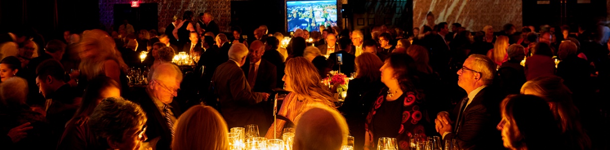 A crowd sitting at tables lit by candlelight which is casting an orange glow on people's faces.