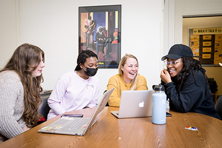 4 female students work on laptops at a table, smiling and laughing.