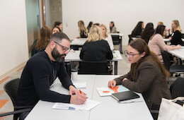 two professionals sitting down and discussing work at a table, group of more people seen together in the background