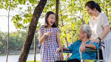 A family including mother, daughter and their grandmother in a wheel chair outside at a lake. They are mid-conversation and laughing