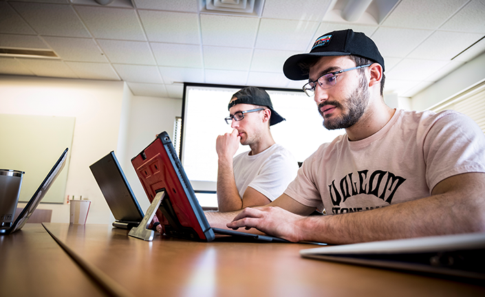 Two students in a side view working on laptops at a desk in classroom. They both have white shirts, black hats and glasses.