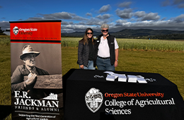 Two people stand behind an Oregon State University College of Agricultural Sciences table at an outdoor event, with an E.R. Jackman Friends and Alumni banner and farmland in the background.