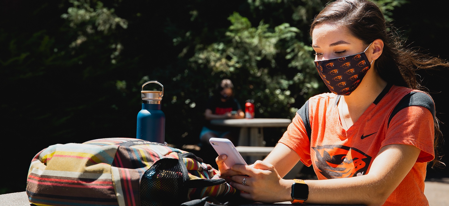 A student in a beaver shirt with a beaver mask looking at her phone at a table.