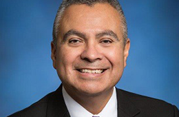 Headshot of Román Hernández. He's smiling with a suit on and there's a blue background.
