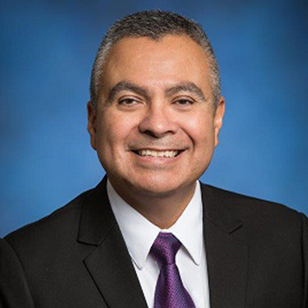 Headshot of Román Hernández. He's smiling with a suit on and there's a blue background.