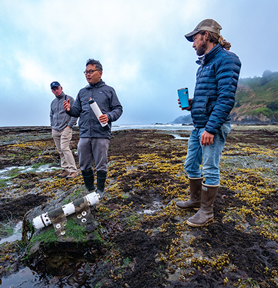 three men standing on rocky ocean shore with scientific equipment