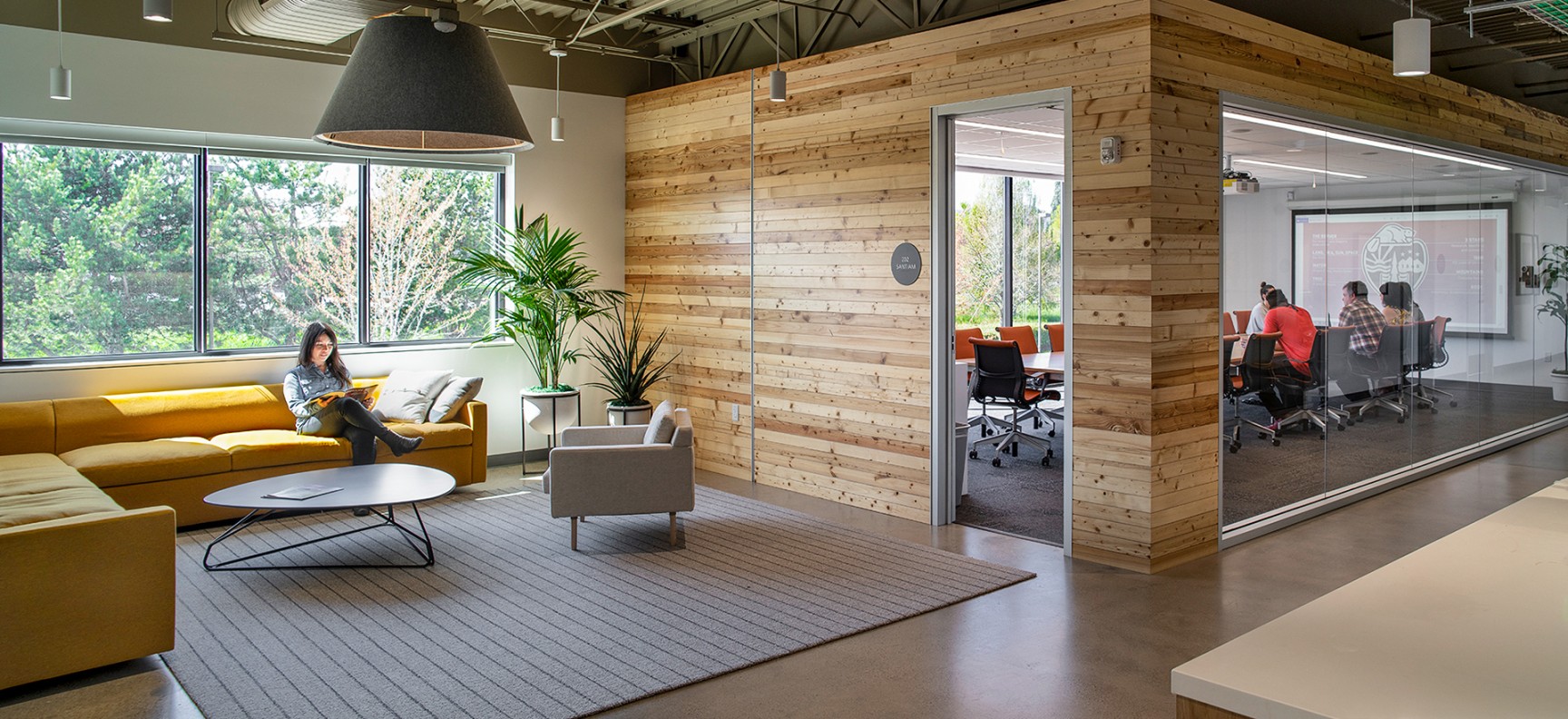 A L-shaped yellow sofa with one person reading on it is on the left side while a meeting room with people are on the right side. The meeting room has a wood panel entrance and one wall made of glass.