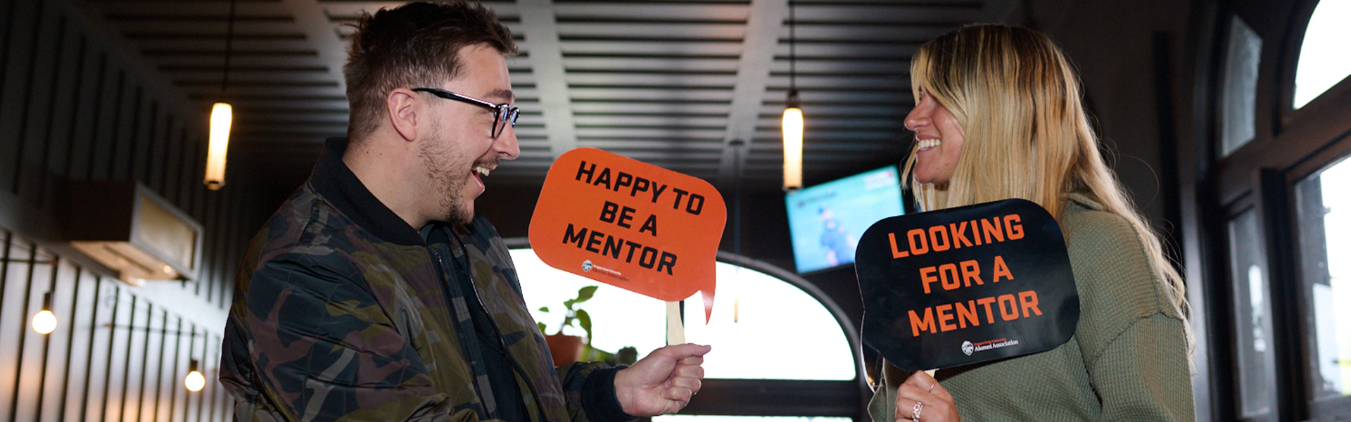 Two people smile and shake hands while holding "Happy to be a mentor" and "Looking for a mentor" signs.