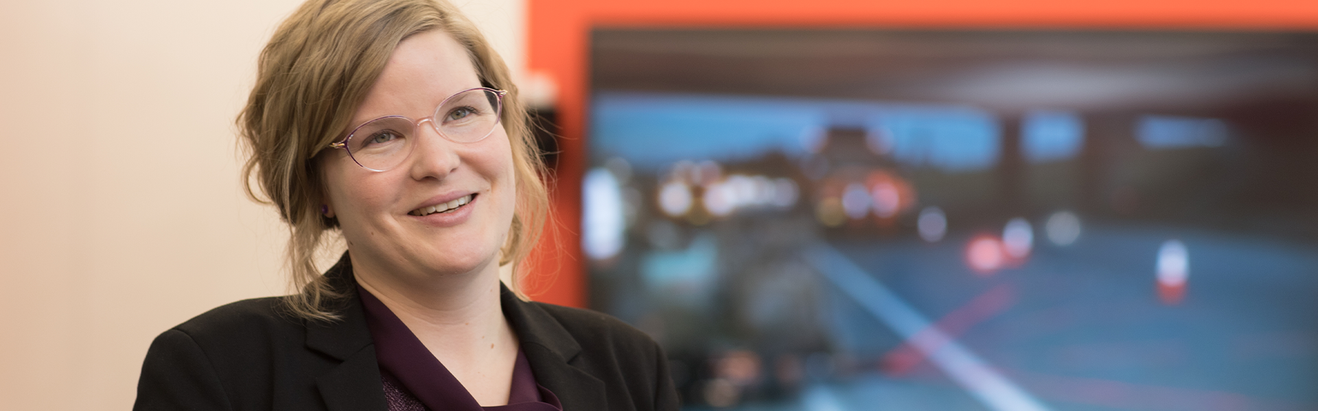 A person in business attire speaks, standing in front of an orange wall and a TV display.