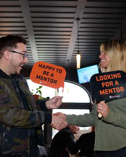 Two people smile and shake hands while holding "Happy to be a mentor" and "Looking for a mentor" signs.