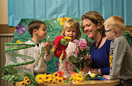 Woman interacting with three children in a learning environment with flowers and hand puppets