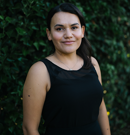 Arlyn Moreno Luna in a black sleeveless dress smiling while standing in front a tall, dark hedge