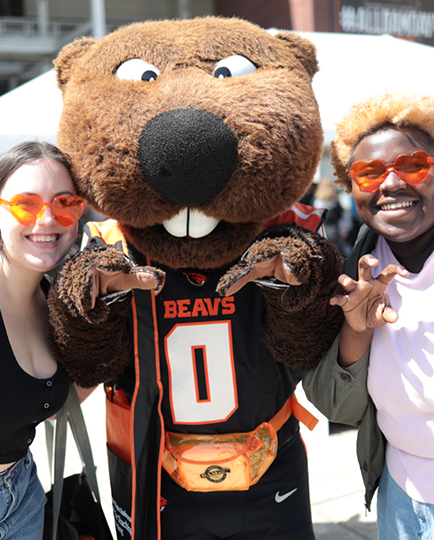 Two students pose with Benny the Beaver while wearing orange heart sunglasses